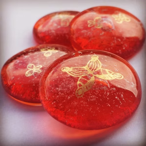 A close-up grouping of red glass cremation stones, each featuring delicate gold butterfly engravings. The stones have a smooth, rounded shape with subtle flecks of cremation ash suspended inside, giving them a soft, glowing appearance.