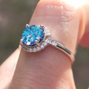 Angled close-up of a sterling silver cremation ring with a round blue dichroic glass centerpiece, secured by prongs and surrounded by clear cubic zirconia accent stones, shown worn on a finger in natural light.