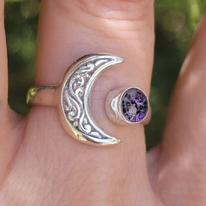 Close-up of a sterling silver crescent moon cremation ring on a finger, showing the detailed engraved moon shape and a round purple crinkle glass stone infused with ashes.