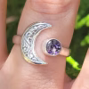 A sterling silver crescent moon cremation ring worn on a finger, featuring an engraved crescent moon on one side and a round purple crinkle glass stone infused with ashes on the other.