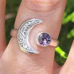 A sterling silver crescent moon cremation ring worn on a finger, featuring an engraved crescent moon on one side and a round purple crinkle glass stone infused with ashes on the other.