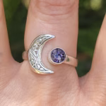 A sterling silver crescent moon cremation ring worn on a finger, featuring an engraved crescent moon on one side and a round purple crinkle glass stone infused with ashes on the other.