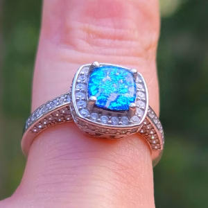 Front-facing close-up of the Cremation Ring with Square Halo Band worn on a finger, showcasing a vivid blue silver crinkle glass center stone set in a square halo of clear cubic zirconia, with a split sterling silver band accented with cubic zirconia stones.