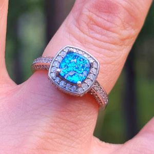 Angled close-up of the Cremation Ring with Square Halo Band worn on a finger, featuring a blue silver crinkle glass center stone secured by four prongs, surrounded by a square halo of clear cubic zirconia and a sterling silver band set with cubic zirconia accents.