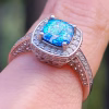 Angled close-up of the Cremation Ring with Square Halo Band worn on a finger, featuring a blue silver crinkle glass center stone secured by four prongs, surrounded by a square halo of cubic zirconia and a split sterling silver band accented with cubic zirconia.