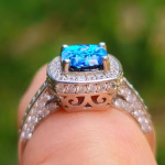 Side-angle close-up of the Cremation Ring with Square Halo Band on a finger, highlighting the blue silver crinkle glass center stone, square halo of cubic zirconia accent stones, decorative gallery details, and a sterling silver band with cubic zirconia accents.