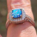 Close-up of the Cremation Ring with Square Halo Band on a finger, showing a blue silver crinkle glass center stone secured by four prongs, surrounded by a square halo of cubic zirconia accent stones on a sterling silver band.