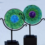 Pair of circular memorial glass suncatchers containing cremation ash, featuring translucent green glass with textured patterns and contrasting blue and purple centers, displayed on black metal stands indoors with a snowy outdoor landscape visible through the window behind them.