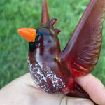Close-up of a handcrafted glass cardinal memorial with cremation ash visible inside the red glass body, held in hand against a grassy background.