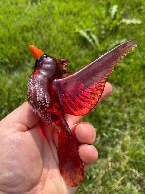 Handheld glass cardinal memorial sculpture in rich red tones, featuring cremation ash suspended within the glass body, photographed outdoors against a green background.