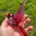 Handheld red glass cardinal memorial sculpture with cremation ash fused into the glass, wings outstretched, shown outdoors over green grass.
