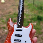 Memorial Guitar with Cremation Ash shown from the front with red glass body, white pickguard, black pickups, and black glass neck with white fret markers, held in hand outdoors