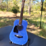 Blue glass memorial guitar sculpture incorporating cremation ash, displayed outdoors on a small stand.