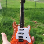 Memorial Guitar with Cremation Ash shown from the front with red glass body, white pickguard, black pickups, and black glass neck with white fret markers, held in hand outdoors