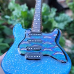 Memorial Guitar with Cremation Ash shown from the front with blue glass body, iridescent wave pattern, black pickups, and white fret markers, held in hand outdoors