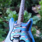 Memorial Guitar with Cremation Ash shown in tie dye blue glass with iridescent details, held in hand outdoors with green garden background