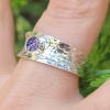 A close-up view of a Hammered Cigar Cremation Ring worn on a finger, featuring a wide sterling silver hammered band with a bezel-set purple crinkle memorial stone at the center.