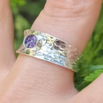 A close-up view of a Hammered Cigar Cremation Ring worn on a finger, featuring a wide sterling silver hammered band with a bezel-set purple crinkle memorial stone at the center.
