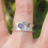 A hammered sterling silver cigar-band ring worn on a finger, featuring a small bezel-set purple crinkle cremation stone centered on the wide textured band.