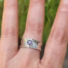 A hand wearing a wide hammered sterling silver cigar-style ring featuring a small bezel-set purple crinkle cremation stone centered on the band.