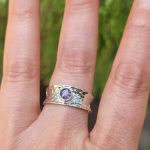 A hand wearing a wide hammered sterling silver cigar-style ring featuring a small bezel-set purple crinkle cremation stone centered on the band.