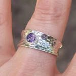 A close-up view of a Hammered Cigar Cremation Ring worn on a finger, featuring a wide sterling silver hammered band with a bezel-set purple crinkle memorial stone at the center.
