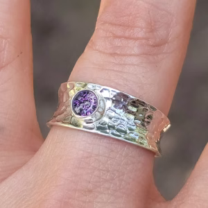 A close-up view of a Hammered Cigar Cremation Ring worn on a finger, featuring a wide sterling silver hammered band with a bezel-set purple crinkle memorial stone at the center.
