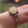 A sterling silver cremation ring featuring a round red orange memorial glass stone infused with cremation ash. The glass displays shifting tones of gold, orange, and deep red with iridescent highlights. The stone is held in a four-prong setting, and the band includes delicate heart-shaped filigree details. The ring is shown on a finger against a softly blurred background with warm bokeh lights.