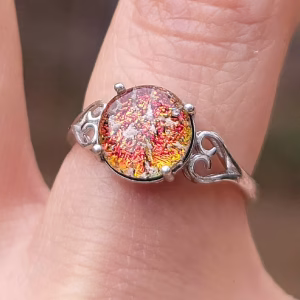 A sterling silver cremation ring featuring a round red-orange memorial glass stone with shimmering embedded ashes. The stone is held by four prongs and framed by delicate heart-shaped filigree on each side. The ring is shown worn on a finger in natural light, highlighting the fiery sparkle of the glass.