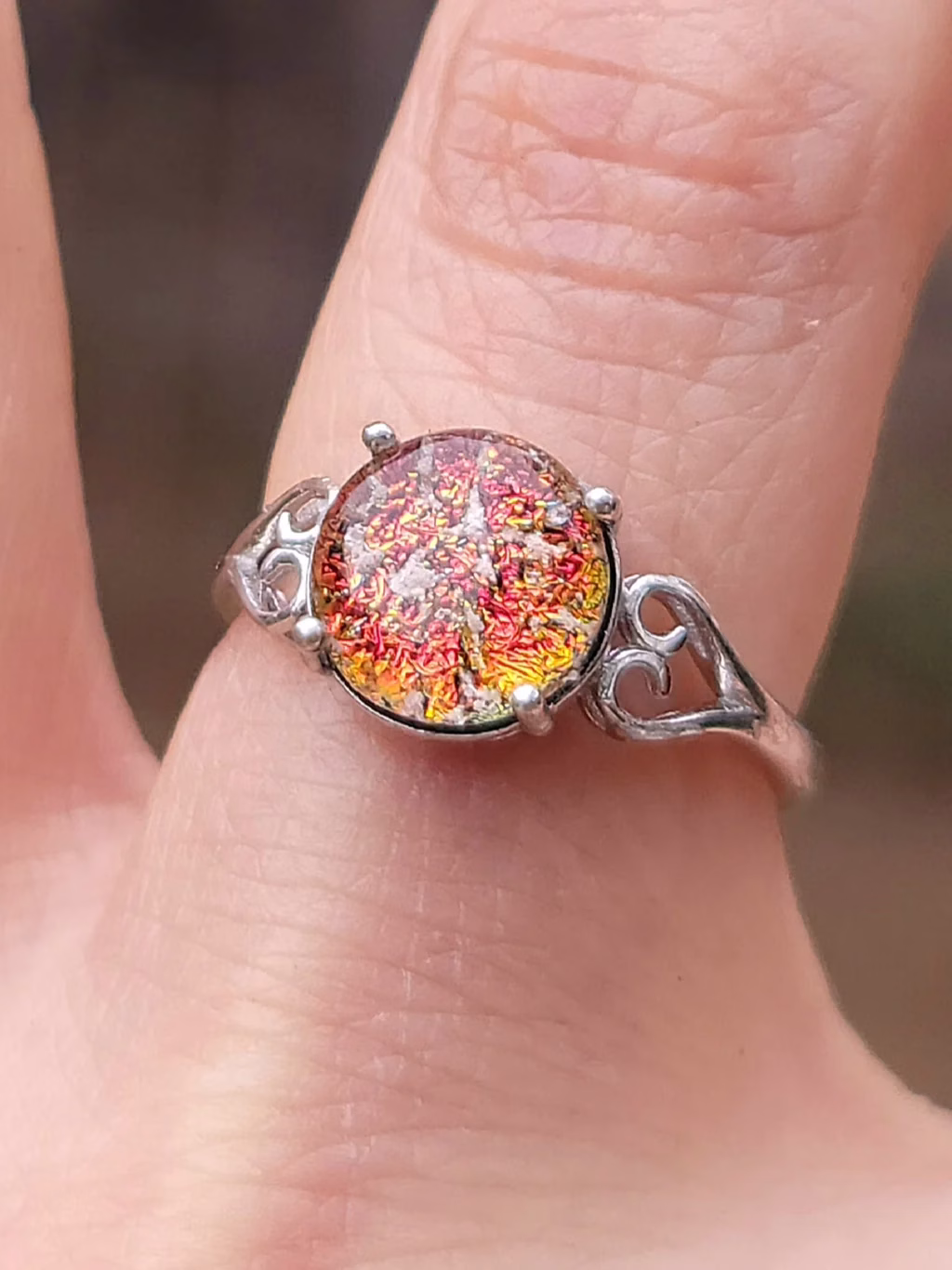 A sterling silver cremation ring featuring a round red-orange memorial glass stone with shimmering embedded ashes. The stone is held by four prongs and framed by delicate heart-shaped filigree on each side. The ring is shown worn on a finger in natural light, highlighting the fiery sparkle of the glass.