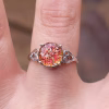 A sterling silver cremation ring featuring a round red-orange memorial glass stone with shimmering embedded ashes. The stone is held by four prongs and framed by delicate heart-shaped filigree on each side. The ring is shown worn on a finger in natural light, highlighting the fiery sparkle of the glass.