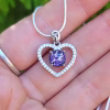 Close-up of a heart-shaped cremation pendant in sterling silver, accented with cubic zirconia stones and featuring a round purple memorial stone, displayed on a necklace resting on a hand.