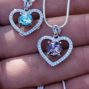 Heart-shaped cremation pendants in sterling silver featuring cubic zirconia accents and round memorial stones, displayed on necklaces resting on a hand.
