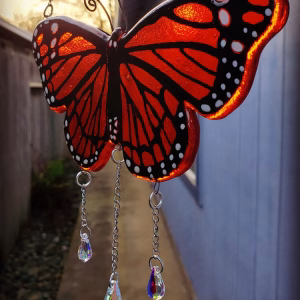 Handcrafted Monarch Butterfly Suncatcher with Cremation Ash, featuring orange and black stained glass butterfly wings with white detailing and three hanging crystal prisms, photographed outdoors in natural light