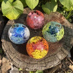 Four spherical Flower Blossom Cremation Paperweights sit on a sunlit tree stump. Each orb contains cremation ashes and vibrant internal floral designs: one in blue and white, one in pink and red, one in green and yellow, and one in orange and yellow. Heart-shaped green leaves surround the stump, adding a natural garden backdrop.