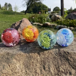 Four spherical glass cremation paperweights arranged on a large rock outdoors. Each paperweight contains a vibrant internal flower design with infused ashes. The colors shown are pink and red, orange and yellow, green and blue, and light blue with white and blue. The background features grass, rocks, and trees under natural sunlight.