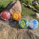 Four round glass cremation paperweights displayed outdoors on a sunlit rock, each featuring a vibrant internal flower design. The colors shown are red and pink, orange and yellow, green and blue, and light blue with white and blue accents. The paperweights have suspended ashes incorporated into the floral patterns.