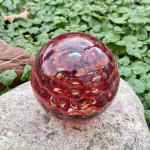 Handcrafted red glass cremation paperweight resting on a natural stone, shown from a top-angled view with layered rippled swirl patterns, clear raised droplets, and warm red and amber tones that highlight the encased ashes and internal depth.