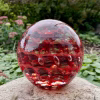 Handcrafted red glass cremation paperweight displayed outdoors on a natural stone, featuring layered rippled swirl patterns with encased ashes subtly visible throughout, highlighted by rich red tones, clear raised droplets, and reflective depth against a garden backdrop.