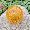 Handcrafted orange glass cremation paperweight shown from a top-down angle, featuring layered rippled swirl patterns with encased cremation ashes throughout, displayed outdoors on a natural stone with green foliage in the background to highlight its vibrant color, depth, and artisan detail