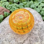 Handcrafted orange glass cremation paperweight shown from a top-down angle, featuring layered rippled swirl patterns with encased cremation ashes throughout, displayed outdoors on a natural stone with green foliage in the background to highlight its vibrant color, depth, and artisan detail