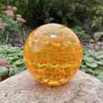 Handcrafted orange glass cremation paperweight featuring vibrant layered rippled swirl patterns with encased cremation ashes throughout, displayed outdoors on a natural stone with soft garden greenery in the background to highlight its color, texture, and artistry