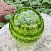 Handcrafted green glass cremation paperweight shown from a top-down angle, featuring layered rippled swirl patterns with encased cremation ashes throughout, displayed outdoors on a natural stone with garden greenery in the background to highlight depth, color, and fine glass artistry