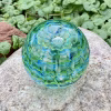 Handcrafted green and blue glass cremation paperweight shown from a top-down angle, featuring layered rippled swirl patterns with encased cremation ashes throughout, displayed outdoors on a natural stone with lush green foliage in the background to highlight depth, color, and artistry
