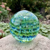 Handcrafted green and blue glass cremation paperweight displayed outdoors on a natural stone, featuring layered rippled swirl patterns with suspended clear droplets and subtle encased ashes, creating an ocean-inspired depth and vibrant color variation.