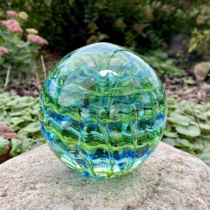 Handcrafted green and blue glass cremation paperweight displayed outdoors on a natural stone, featuring layered rippled swirl patterns with suspended clear droplets and subtle encased ashes, creating an ocean-inspired depth and vibrant color variation.