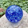 Handcrafted blue glass cremation paperweight shown from a top-down angle, featuring layered rippled swirl patterns in multiple shades of blue with encased cremation ashes throughout, displayed outdoors on a natural stone with green foliage in the background to highlight depth, color, and artistry