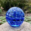 Handcrafted glass cremation paperweight featuring vibrant blue rippled swirl patterns with encased cremation ashes throughout, displayed outdoors on a natural stone with soft garden greenery in the background to highlight color, texture, and artistry
