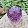 Handcrafted glass cremation paperweight shown from a top-down angle, featuring flowing pink and purple rippled swirl patterns with encased cremation ashes throughout, displayed outdoors on a natural stone with lush green foliage in the background