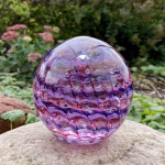 Handcrafted glass cremation paperweight featuring pink, purple, and clear rippled swirl patterns with encased cremation ashes, displayed outdoors on a natural stone with garden greenery in the background to highlight color, texture, and artistry